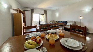 Un'area pranzo con un tavolo di legno apparecchiato per la colazione, con piatti di frutta e succo, e sullo sfondo si intravede un soggiorno con divano e poltrone.