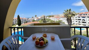 Un balcone con un tavolo apparecchiato per la colazione, con frutta e bevande, affacciato su una vista urbana che include edifici e una piscina.