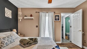 A bedroom with a large bed, a bedside table with a lamp, a wall-mounted shelf, and a door leading to another room.