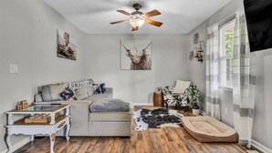 A cozy living room with a ceiling fan, a sofa, a coffee table, a dog bed, and a window with curtains.