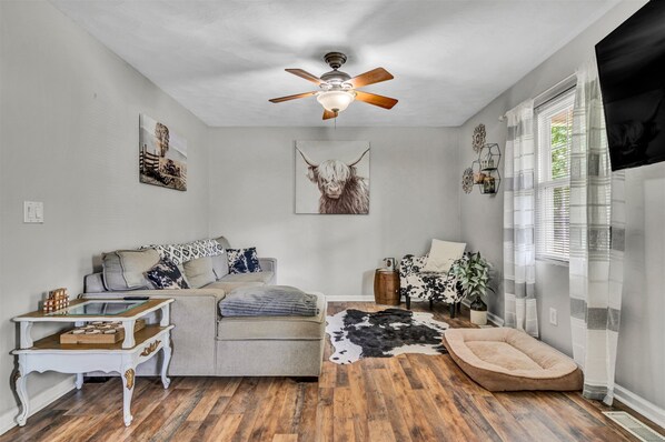 A cozy living room with a ceiling fan, a sofa, a coffee table, a dog bed, and a window with curtains.
