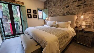 A bedroom with a stone wall, a bed with white bedding, a bench, and a view through a window.
