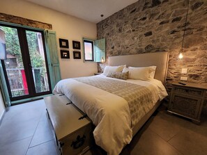 Quarto com parede de pedra, cama com roupa de cama branca, um banco e vista pela janela.
