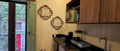 A kitchen with a sink, countertop, and cabinets. There is a trash can and a window with a view of greenery.