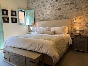 Une chambre avec un mur en pierre, un lit avec une literie blanche, une table de chevet et une commode ornée de motifs décoratifs.