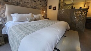 Une chambre avec un mur en pierre, un lit avec une literie blanche, une table de chevet et un miroir agrémenté d’un panneau décoratif.