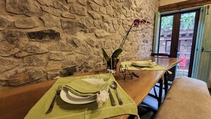 Une table à manger est dressée avec des sets de table verts, des assiettes, des couverts et un vase orné d’une orchidée. La table est placée contre un mur de pierre, offrant une vue sur la verdure à l’extérieur.