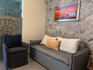 Un mur en pierre orné d’un tableau encadré, un canapé gris avec des coussins et un fauteuil sombre.