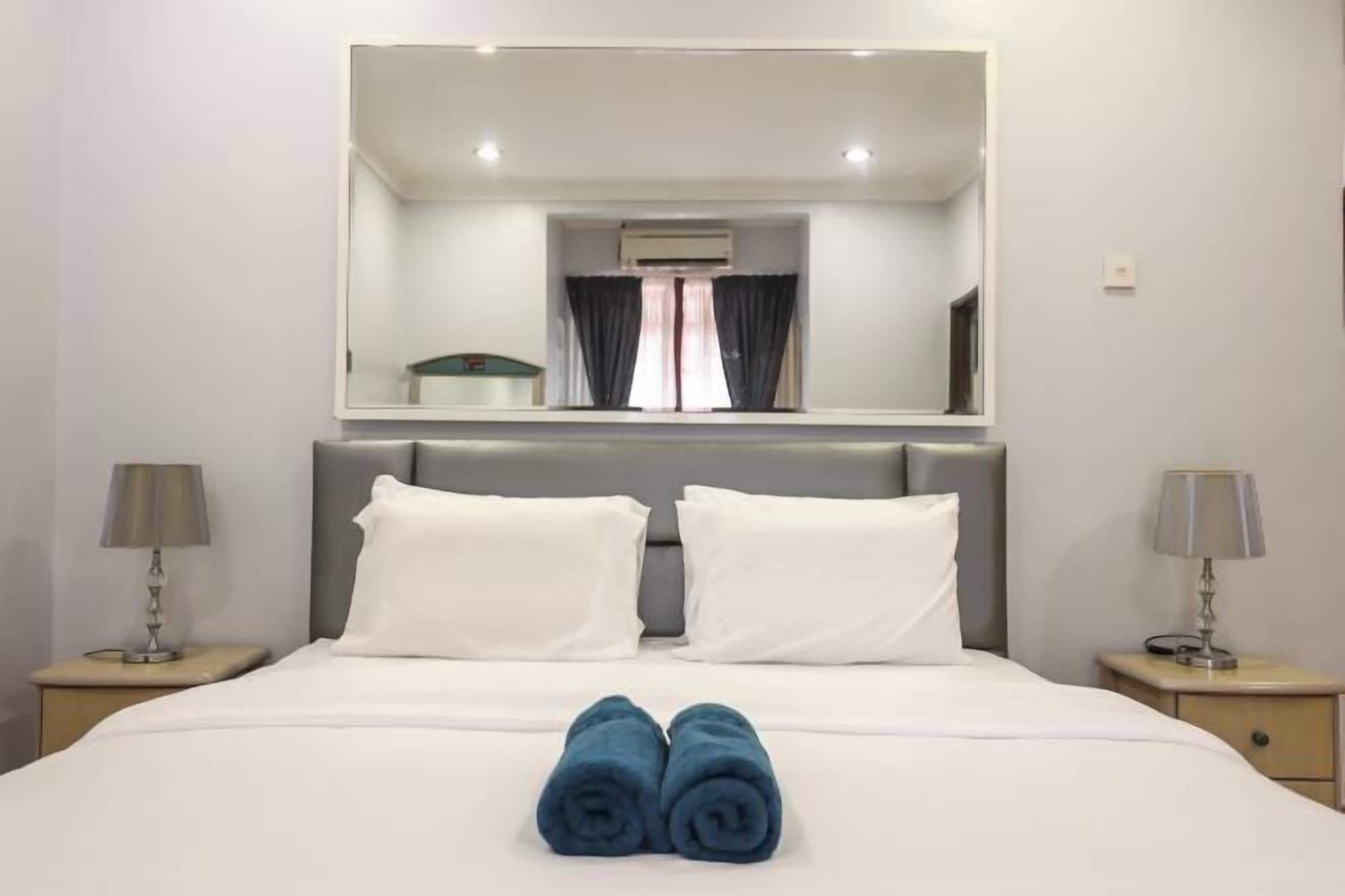 Une chambre d’hôtel avec un grand lit, deux oreillers blancs, une tête de lit, une table de chevet avec une lampe, un miroir au-dessus du lit et une fenêtre avec des rideaux.