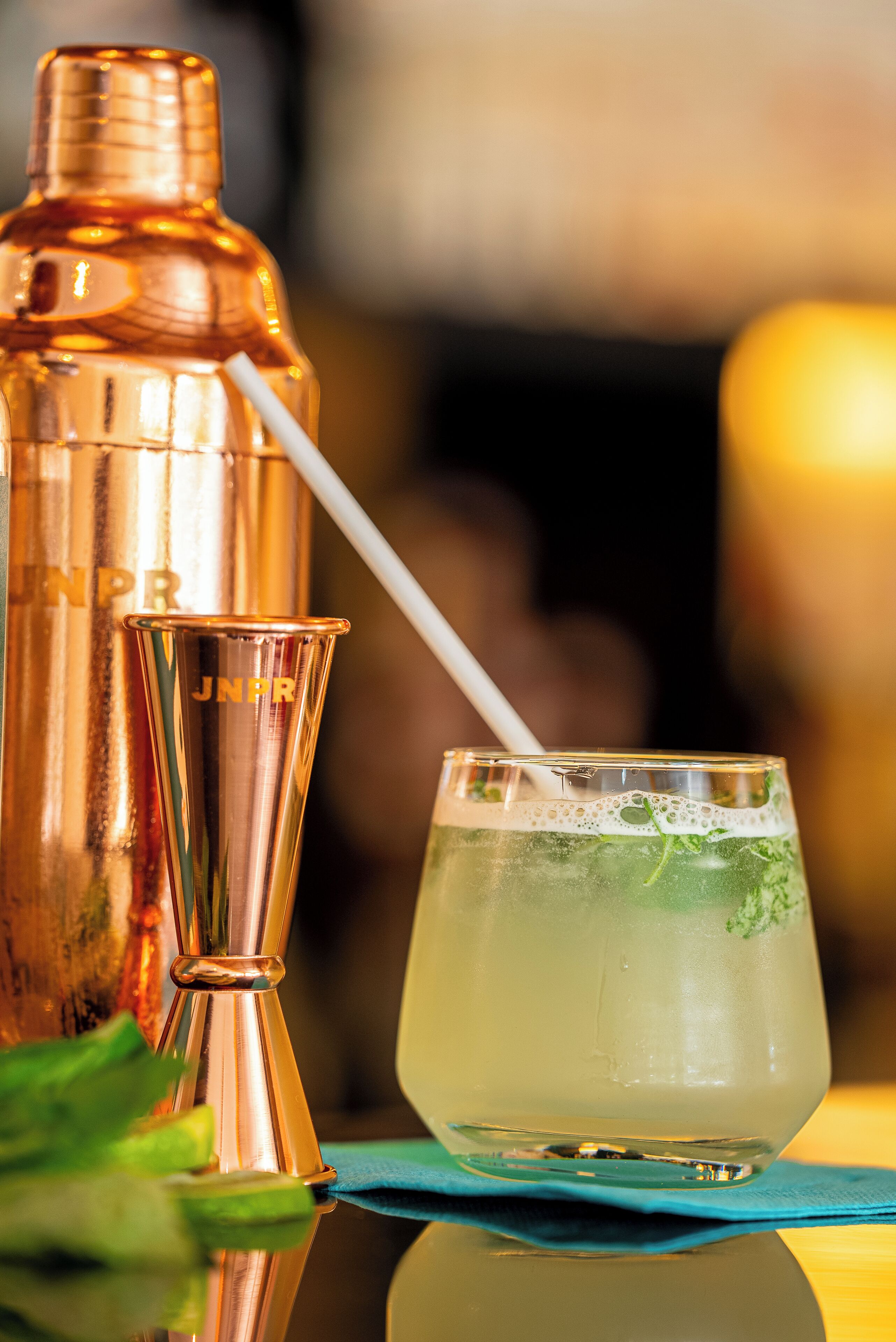 Ein Cocktail mit Strohhalm, ein Messing-Cocktailshaker und ein Glas auf einer spiegelnden Oberfläche.