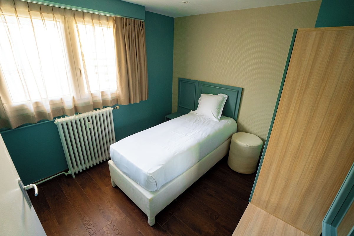 A single-bed room with a wooden floor, a white bed, a headboard, a bedside table, and a radiator.