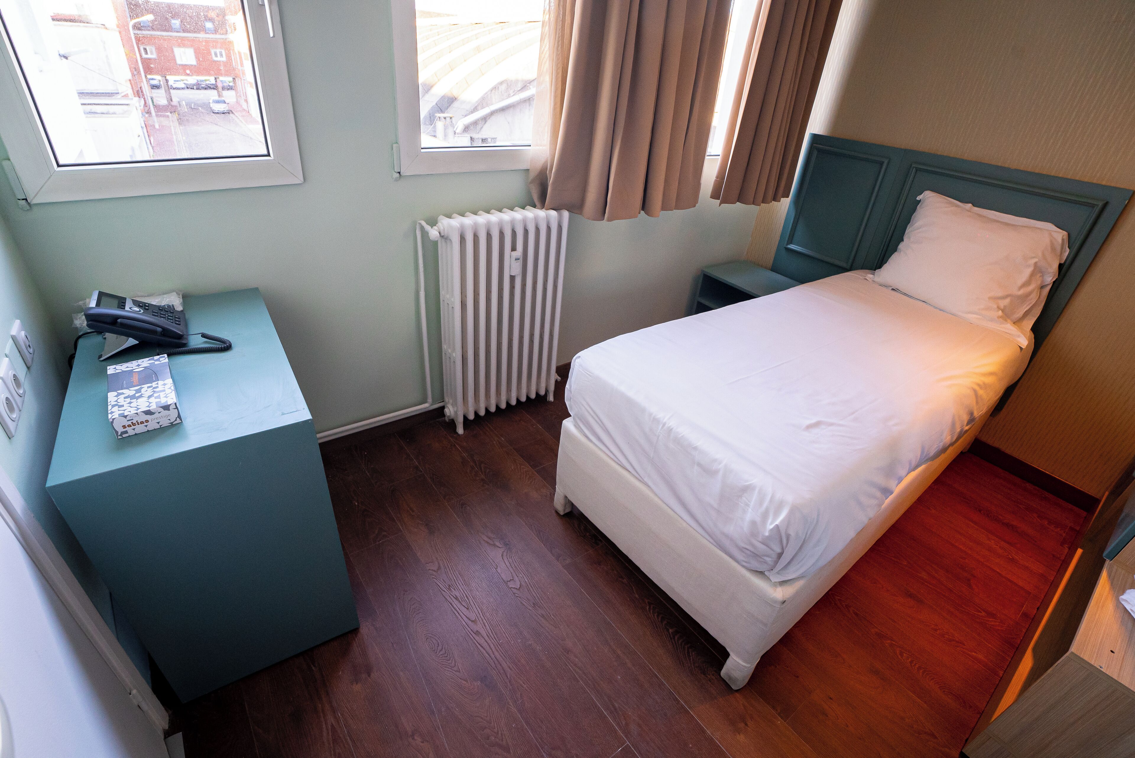 Une chambre d’hôtel avec un lit, une table de chevet avec un téléphone, un radiateur et une fenêtre avec des rideaux.