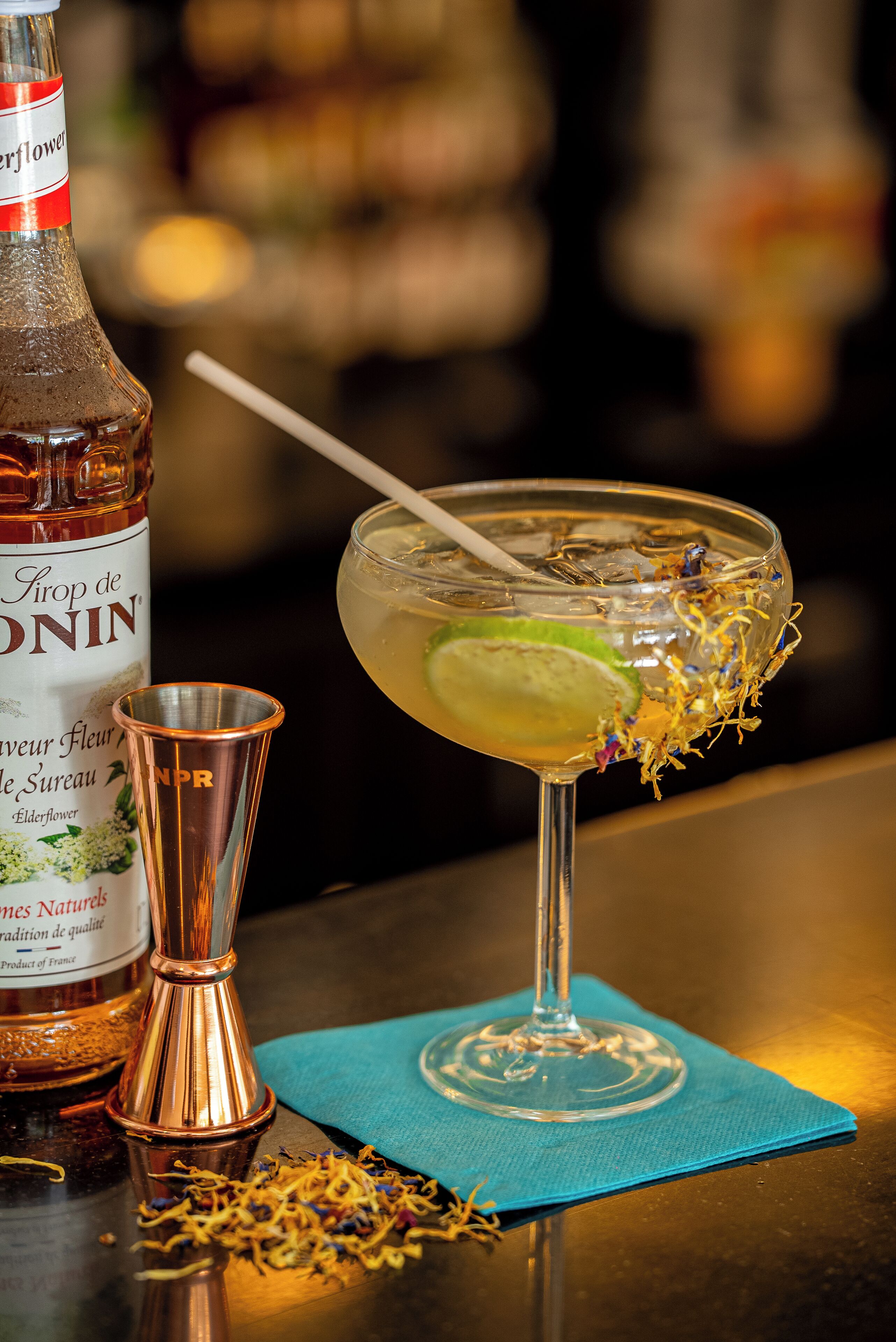 A bottle of elderflower liqueur, a cocktail glass with a lime garnish, and a copper cocktail shaker on a bar counter.