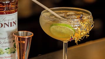 A bottle of elderflower liqueur, a cocktail glass with a lime garnish, and a copper cocktail shaker on a bar counter.