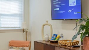 Una sala de estar con un televisor montado en la pared, un mueble de madera, una silla y una mesita con un cuenco y una planta.