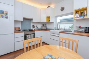 A modern kitchen with white cabinets, a wooden dining table, and a window overlooking the street. - 2-Bed Apartment Steps from Muswell Hill (Greater London)