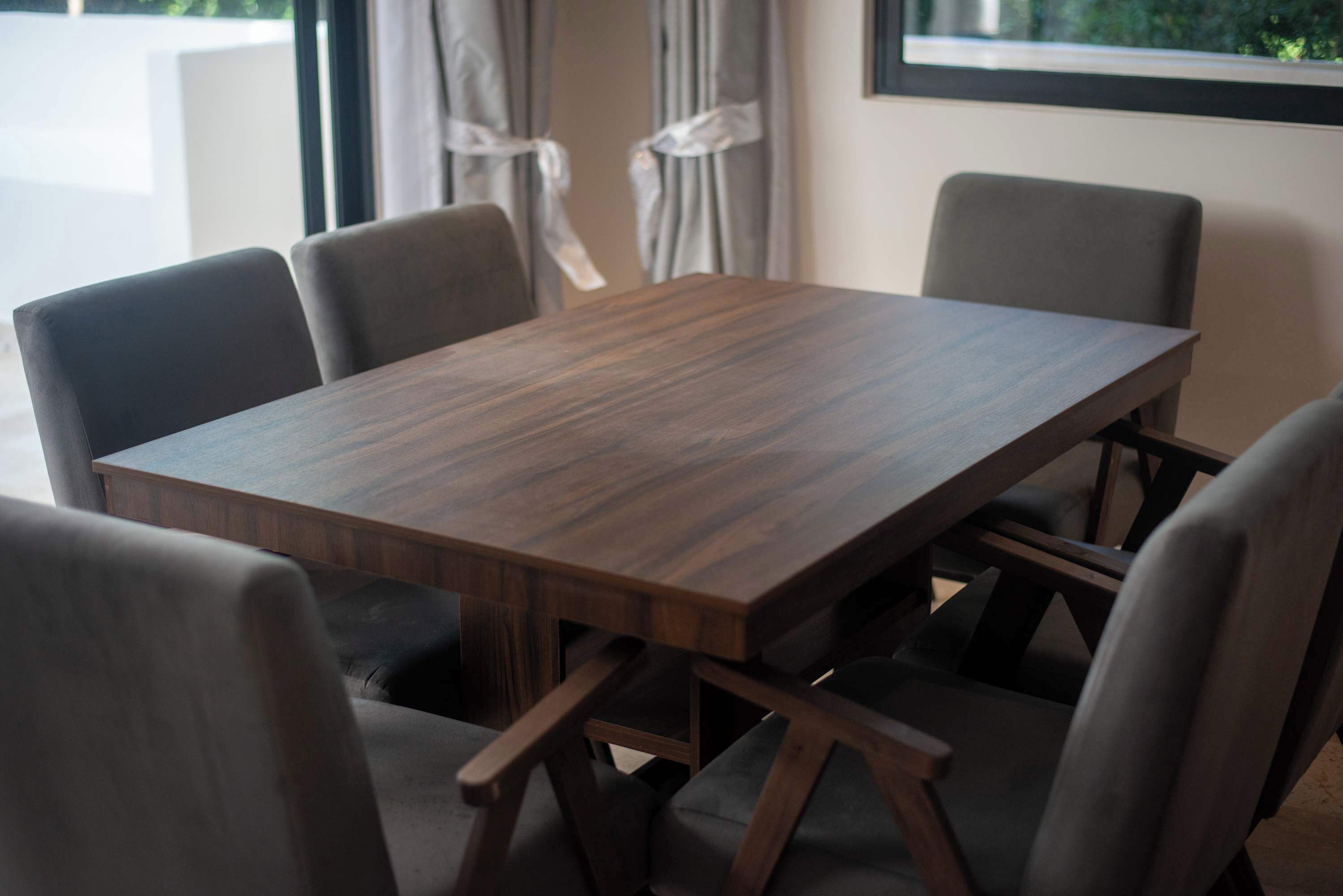 Mesa retangular de madeira com quatro cadeiras, cada uma com almofada escura. A mesa está colocada em um quarto com janela e cortinas.