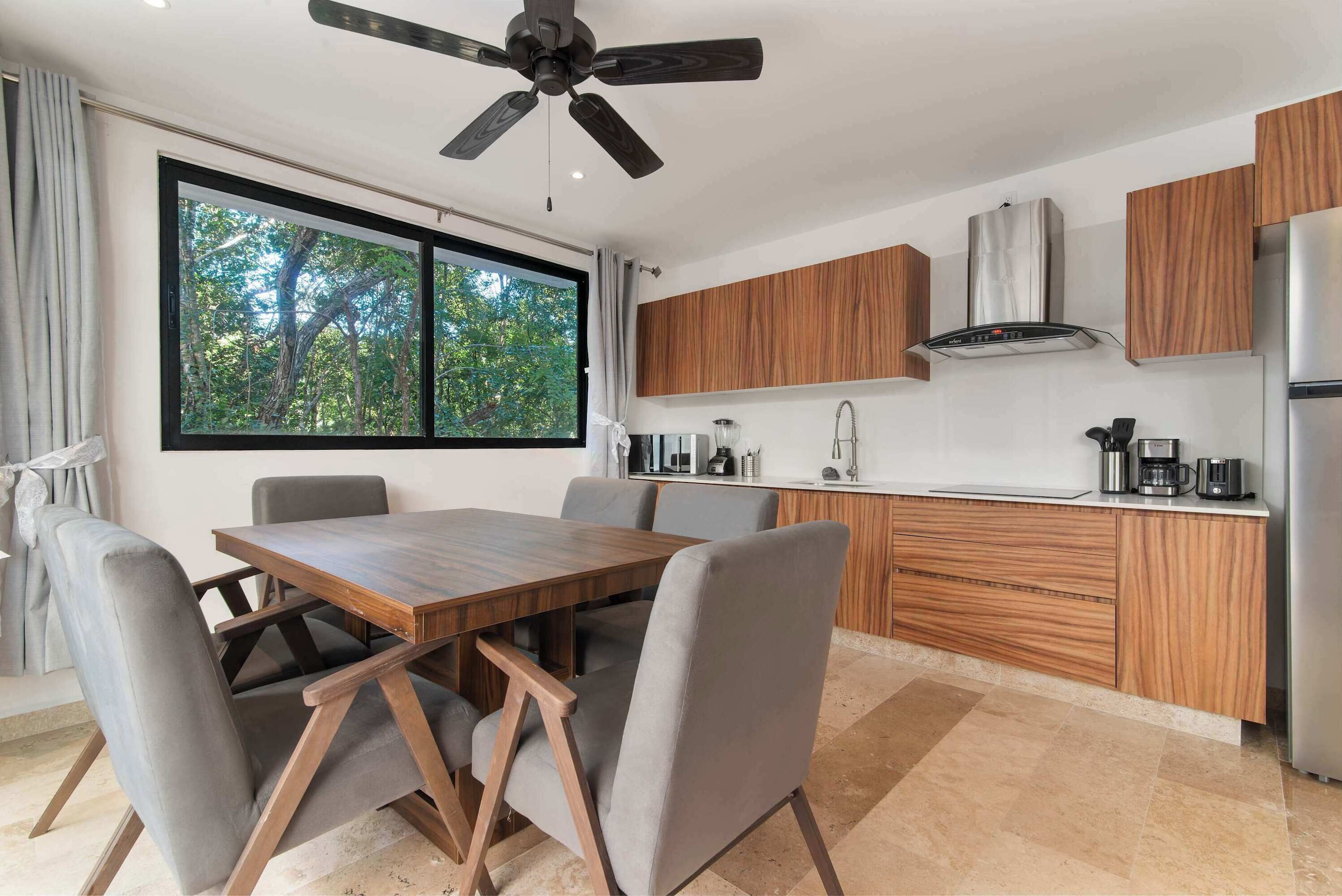 A modern kitchen with a dining table, grey upholstered chairs, wooden cabinets, a stainless steel range hood, and a large window with a view of trees.