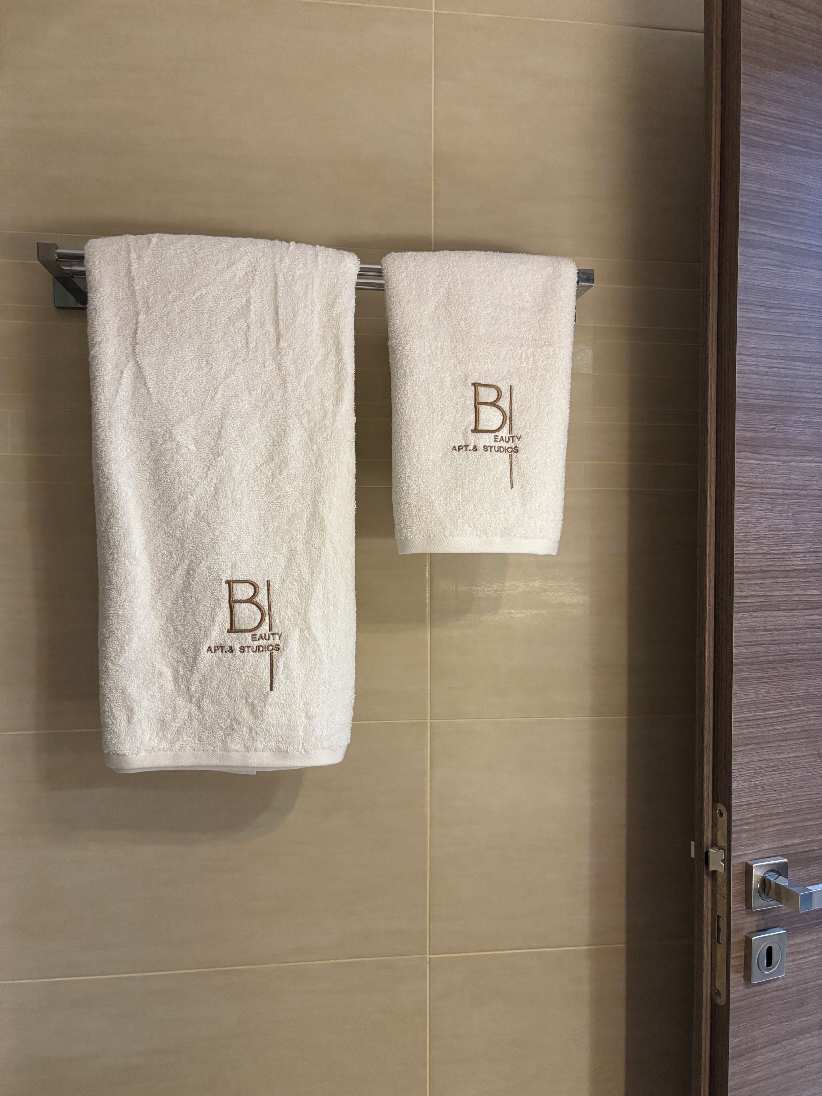 Kaksi valkoista pyyhettä on ripustettu pyyhetelineeseen, ja niissä on painettuna logo, jossa lukee ”BI” ja ”Eauity Apt. & Studios”.