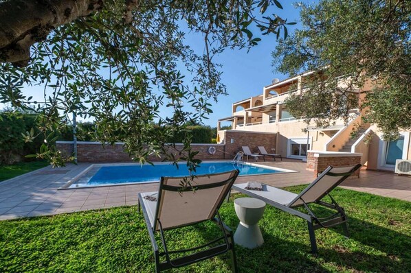 A pool area with two lounge chairs and a table, surrounded by a well-maintained lawn and a building with a modern architectural style.