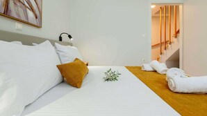 Habitación de hotel con cama, ropa blanca, almohada marrón, una planta pequeña y toallas enrolladas en el suelo.