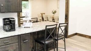 Une cuisine avec des meubles en bois foncé, un plan de travail en marbre et une machine à café. On y trouve également un coin repas avec des chaises blanches et une table en arrière-plan.