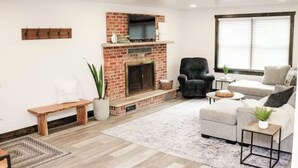 Un salon avec une cheminée en briques, un fauteuil noir, un canapé gris et une grande fenêtre avec des stores.