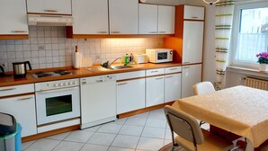 A kitchen with white cabinets, a microwave, a stove, and a sink. There is a dining table with a chair and a window with curtains. - For companies only! Comfortable apartment for fitters near Gießen with own entrance. (Reiskirchen)