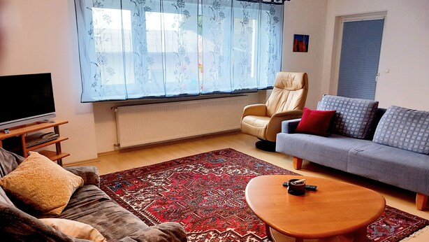 A living room with a sofa, armchair, and a coffee table. There is a TV on a stand, a window with curtains, and a radiator. - For companies only! Comfortable apartment for fitters near Gießen with own entrance. (Reiskirchen)