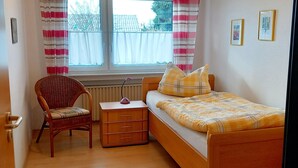 A bedroom with a wooden bed, a wicker chair, a bedside table, and a window with curtains. - For companies only! Comfortable apartment for fitters near Gießen with own entrance. (Reiskirchen)