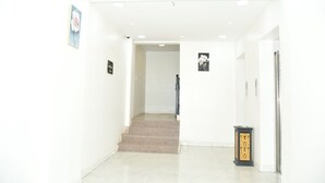 Un pasillo luminoso y blanco con una escalera de mármol que conduce a una puerta. Hay dos cuadros en las paredes y un objeto decorativo negro y dorado en el suelo.