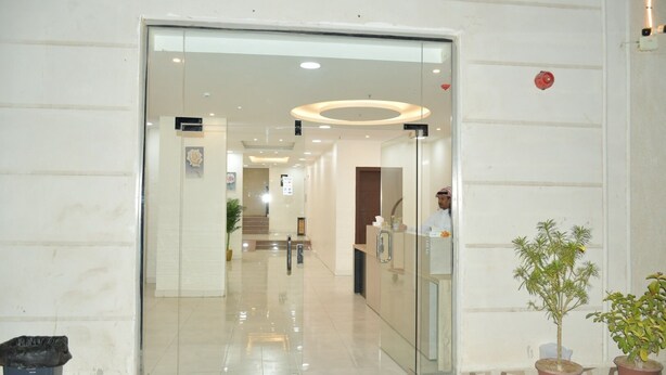 Zona de recepción de un hotel con puerta de cristal, mostrador de recepción y escalera que sube a los pisos superiores.