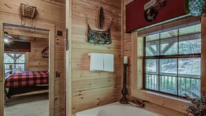 A wooden cabin interior with a large window, a bathtub, a bed with a red and black checkered blanket, and a wall-mounted fish-themed bag. - Amazing 4BR Cabin -HT, Fire Pit & Game Room-10 Mins from PF (Sevierville)