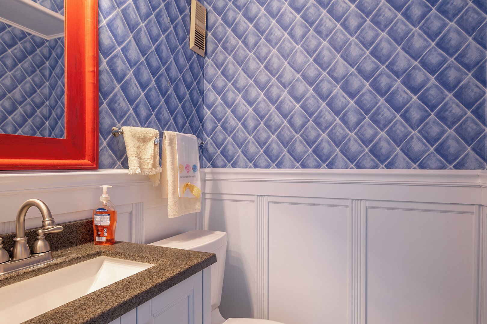 A bathroom with a blue diamond-patterned wall, a white toilet, a granite countertop, a red-framed mirror, and a bottle of hand soap.