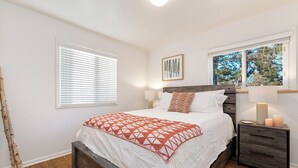 Une chambre à coucher avec un lit en bois, une literie blanche, une table de chevet avec une lampe et une fenêtre avec des stores.