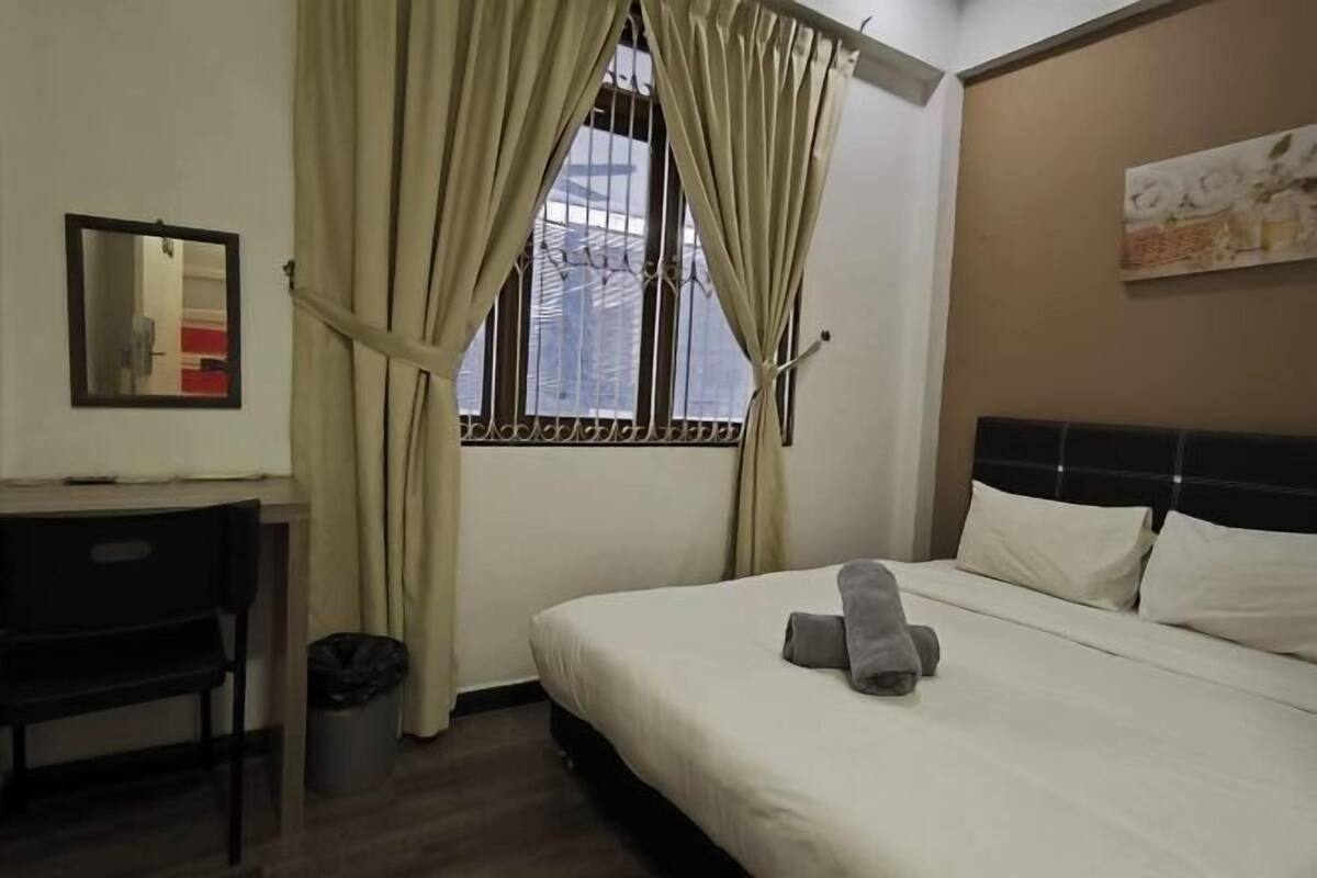 A hotel room with a bed, a desk, a chair, a trash bin, and a window with curtains.