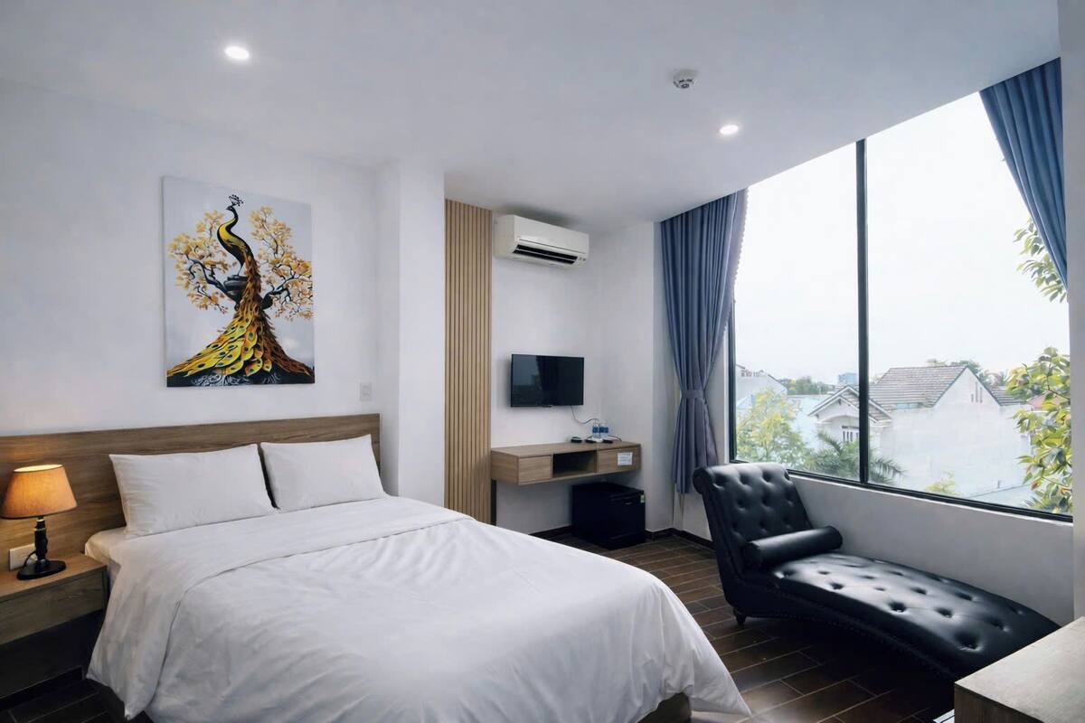 A hotel room with a bed, a desk with a TV, a chair, and a large window with a view of buildings.