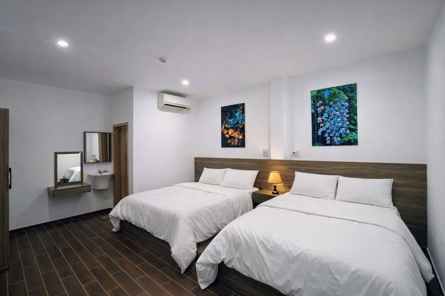 Quarto de hotel com duas camas, uma mesinha de cabeceira com abajur, um espelho, uma pia e dois quadros pendurados na parede.