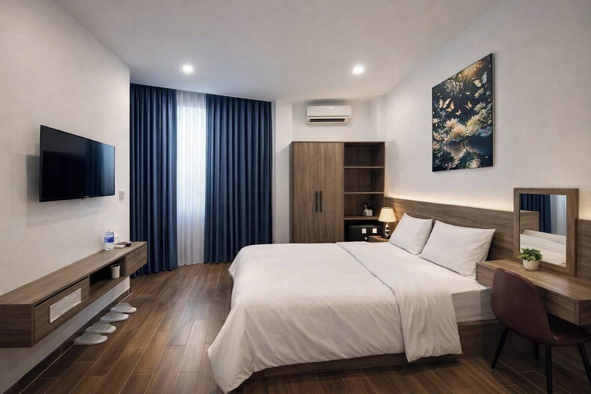 A modern hotel room with a large bed, a flat-screen TV, a wooden wardrobe, a desk with a chair, and a large window with curtains.