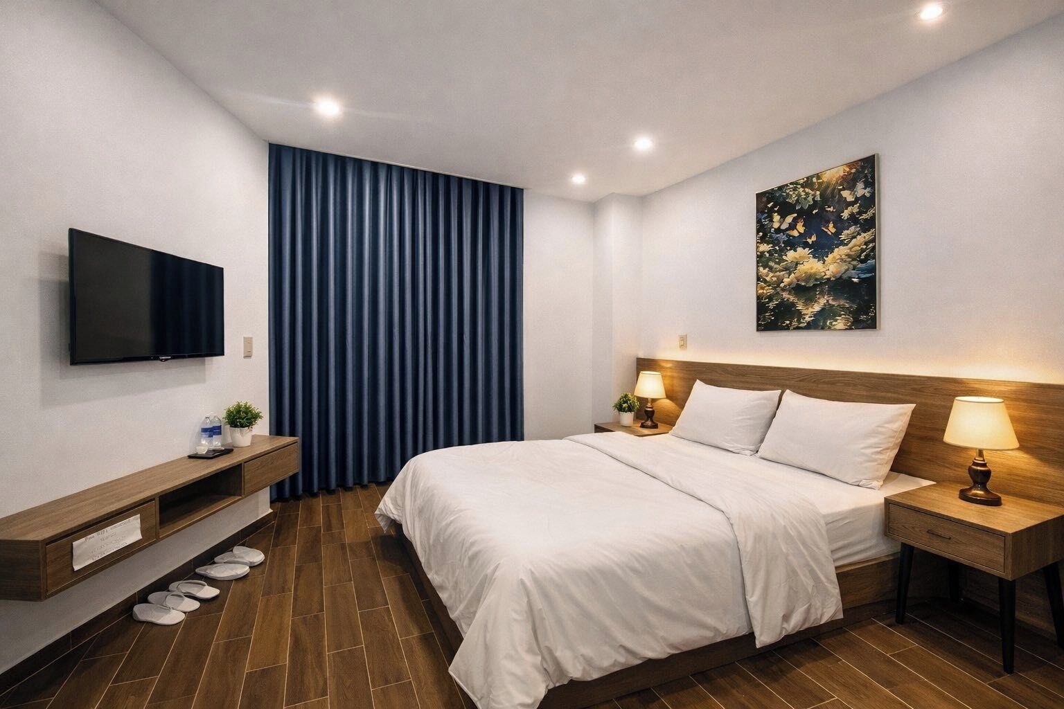 A modern hotel room with a large bed, a flat-screen TV, a wooden nightstand, a bedside table with a lamp, and a blue curtain.