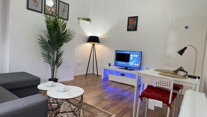 A modern living room with a flat-screen TV, a standing lamp, a potted plant, and a small table with cups.