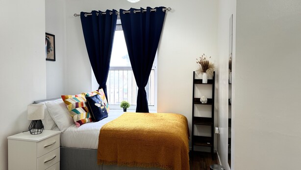 A bedroom with a bed, a nightstand, a shelf with decorative items, and a window with curtains.