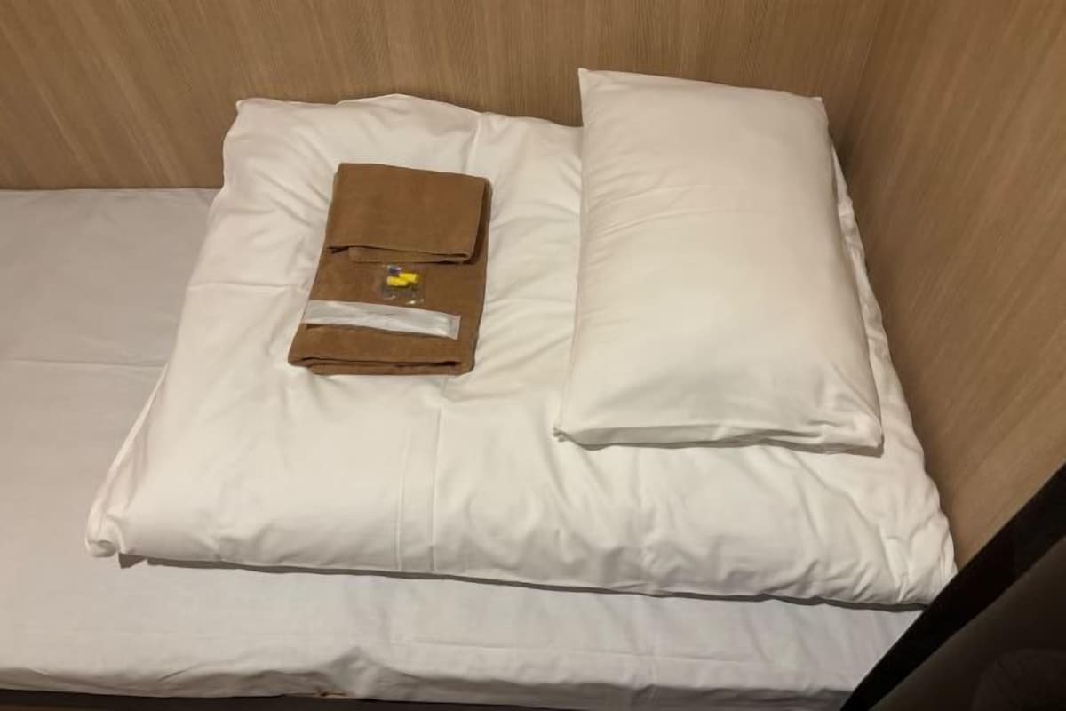 Una cama bien hecha con sábanas y almohadas blancas, una toalla marrón doblada y un objeto pequeño sobre la almohada.