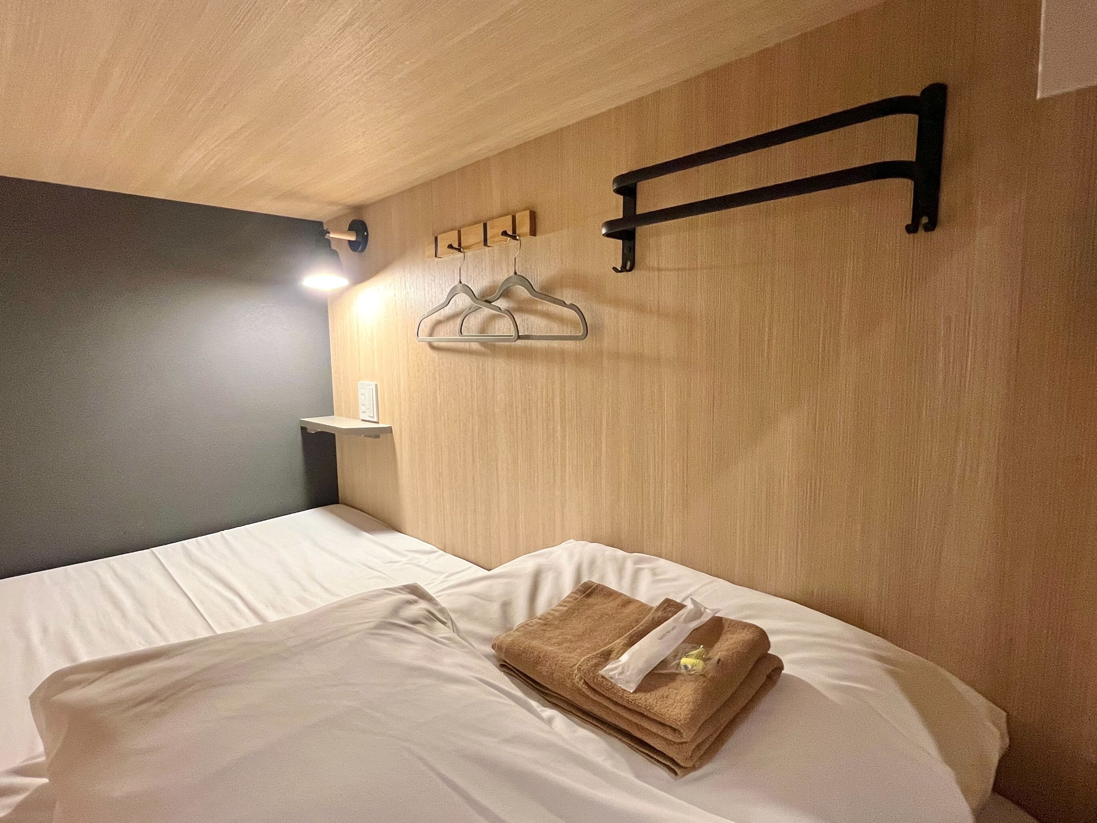 A hotel room with a bed, a wooden headboard, a shelf with a lamp, and a wall-mounted hanger.