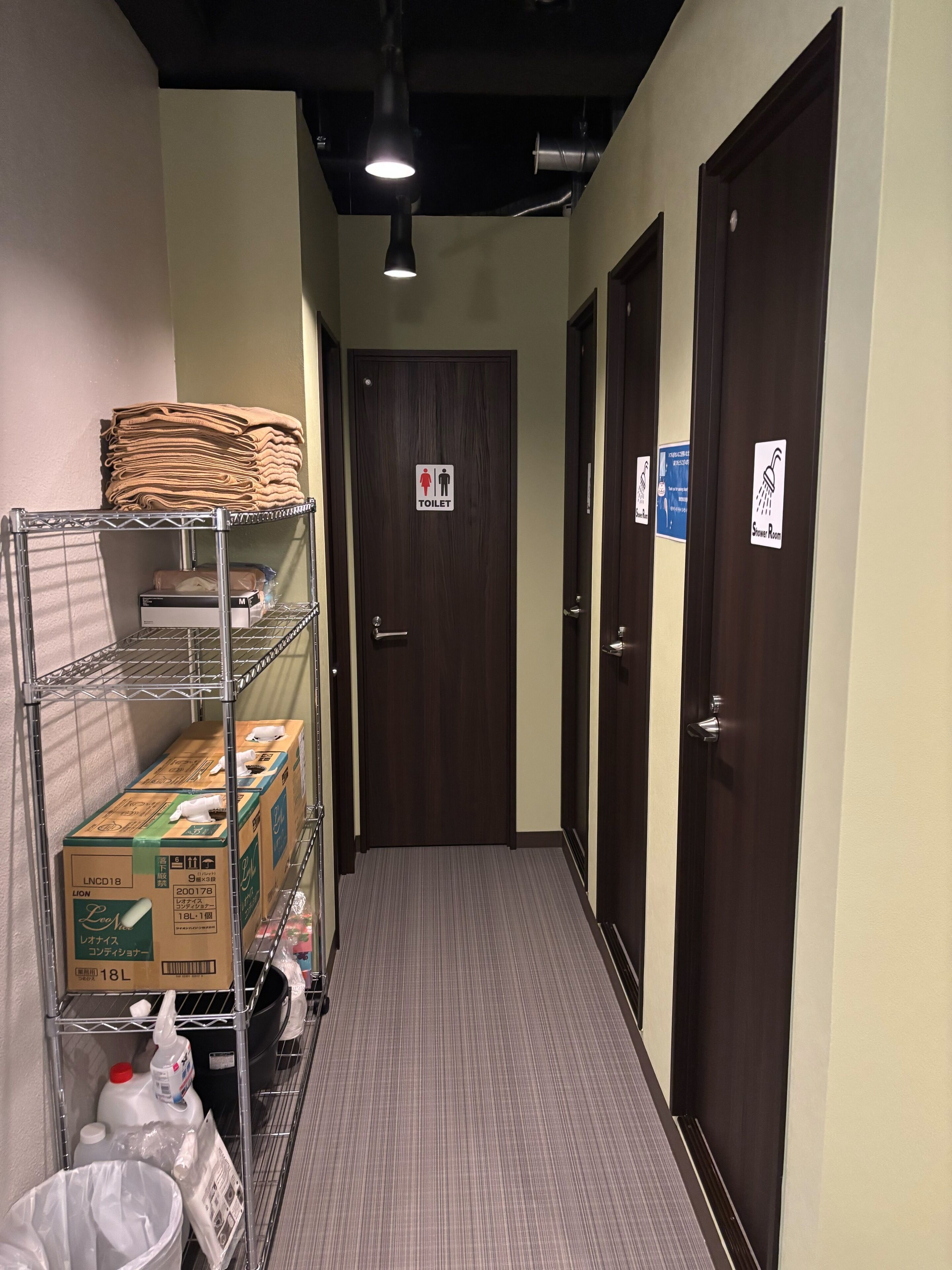 A hallway with a metal rack holding folded towels and boxes, a door with a toilet sign, and a shower sign on adjacent doors.