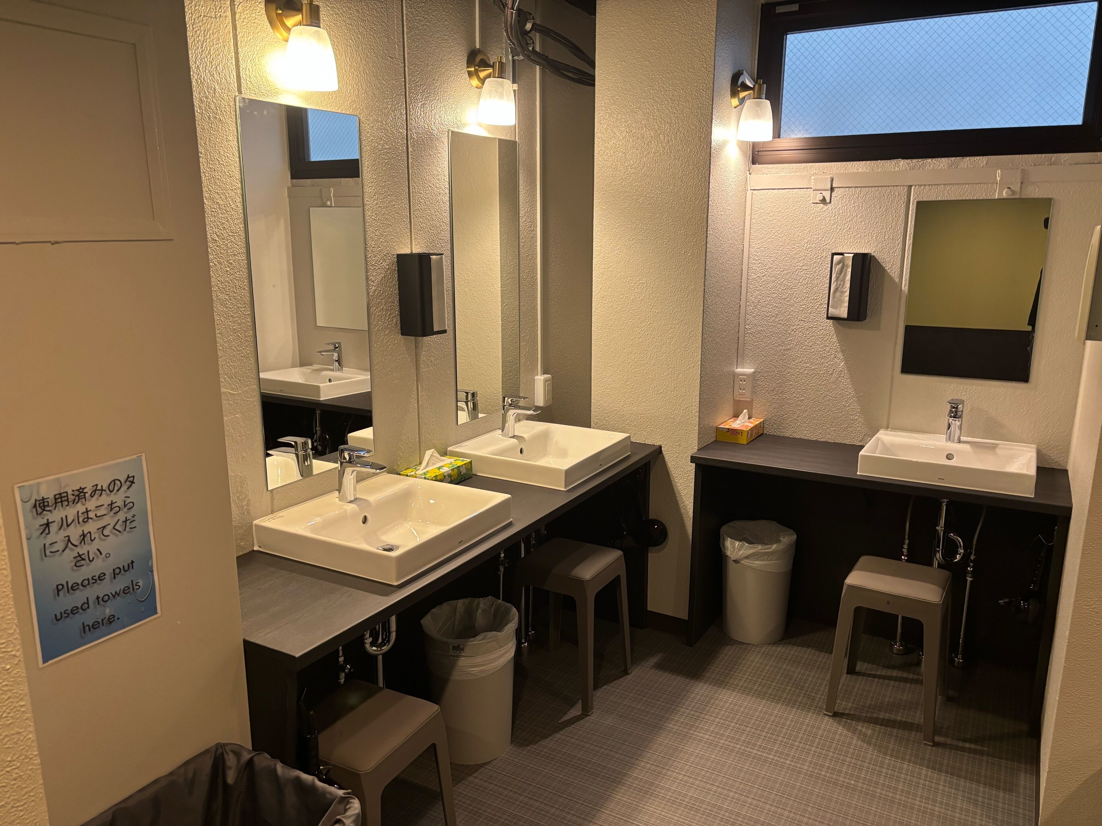 A modern bathroom with two sinks, mirrors, and a trash bin. There is a sign instructing to put used towels there.