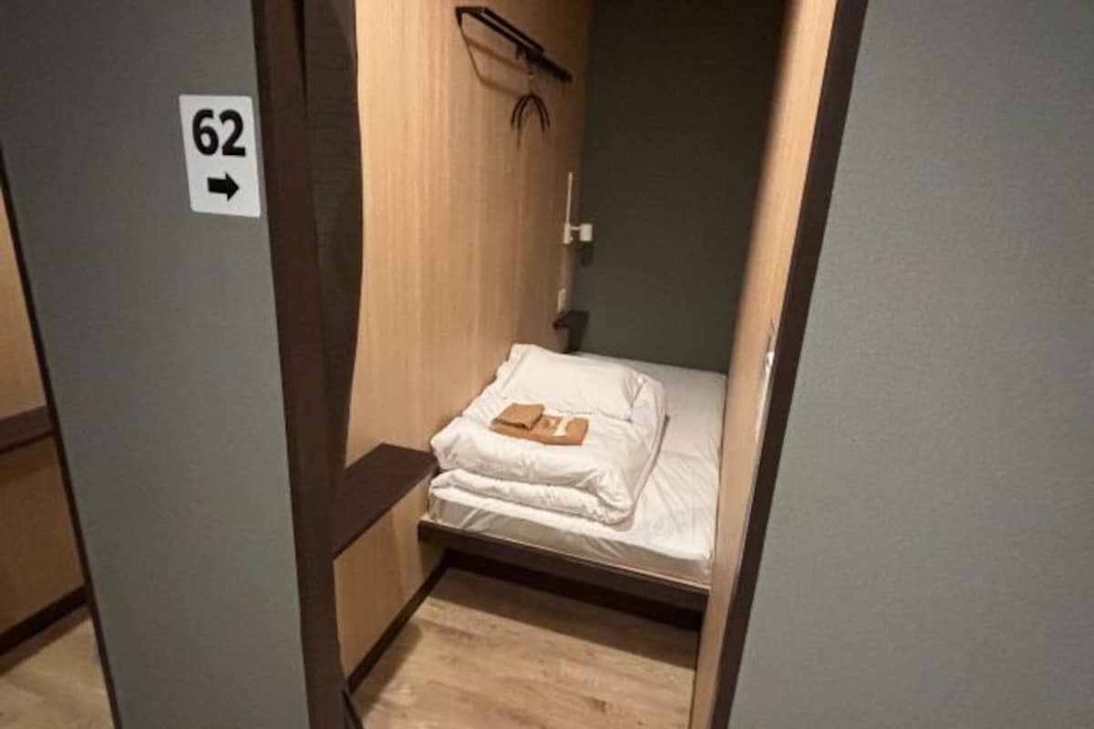 A small, narrow room with a bed, a small table, and a door marked with the number 62.