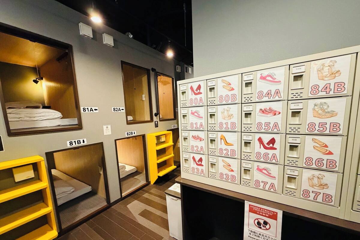 A hotel room with a bed, a yellow bedside table, a mirror, and a shoe rack with labeled shoe boxes.