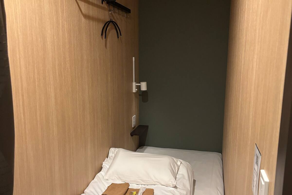 A small, narrow hotel room with a single bed, a small table, a wall-mounted shelf, and a towel rack.