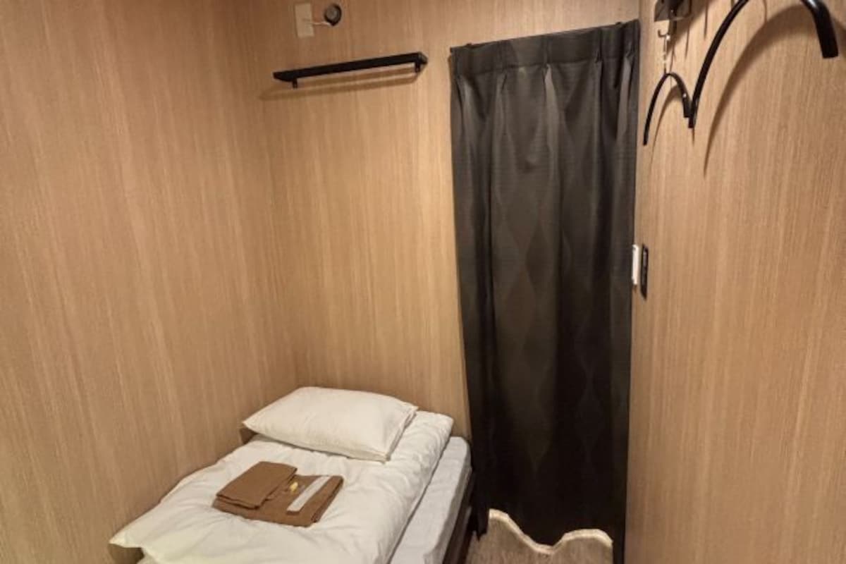 Una habitación pequeña con cama individual, pared de madera, cortina negra y una pequeña estantería sobre la cama.